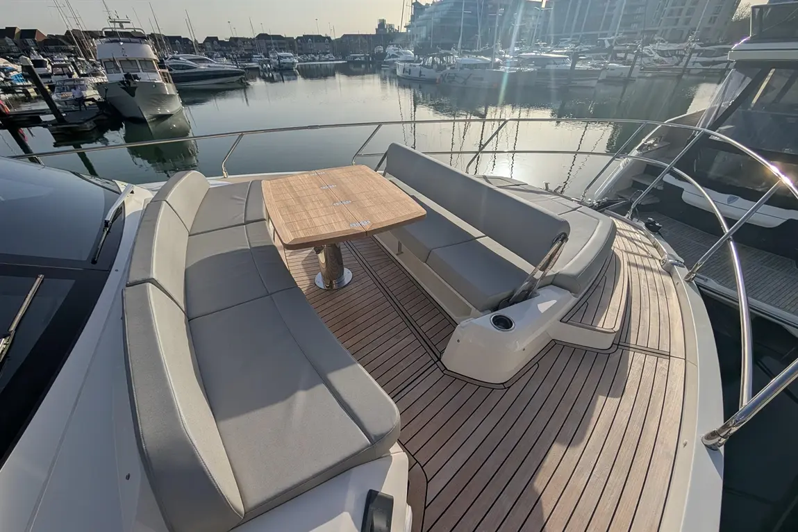 Yacht deck with grey seating, a wooden table, and a view of the marina and boats reflected in calm water.